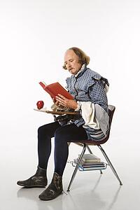 William Shakespeare in period clothing sitting in school desk reading a book.