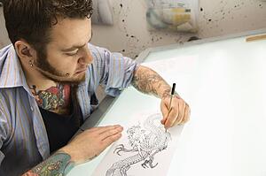 Caucasian male tattoo artist drawing tattoo on light table.
