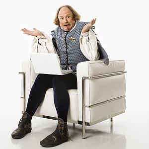 William Shakespeare in period clothing sitting on modern chair with laptop and shrugging with confusion at viewer.