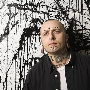 Tattooed and pierced man standing against paint splattered background.