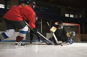 Female hockey player trying to make goal as goalie protects the net.