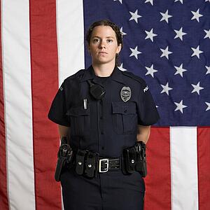 Portrait of mid adult Caucasian policewoman standing with arms behind back and American flag as backdrop.