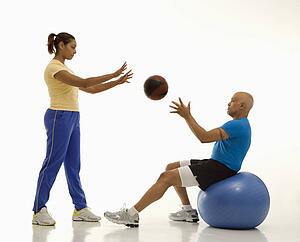 Mid adult woman throwing ball to mid adult man balancing on blue exercise ball