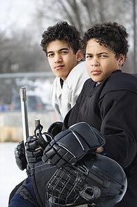 Two boys in ice hockey uniforms sitting on ice rink sidelines looking