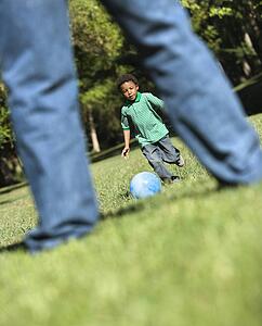 Son running and kicking ball towards father in park.