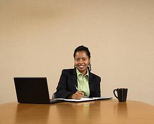 Businesswoman sitting at conference table with laptop computer and coffee cup smiling and writing in book