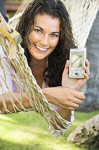 Smiling pretty young adult brunette female laying in hammock holding up PDA towards viewer