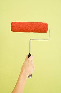 Caucasian female hand holding paint roller dipped in red against green wall.