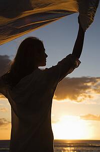 Attractive mid-adult woman holding up fabric in breeze silhouetted by sunset beside ocean