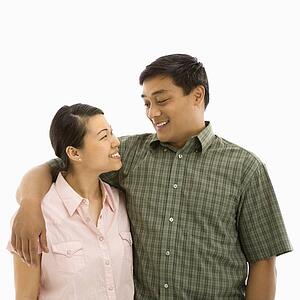 Mid adult Asian couple standing with arms around eachother and smiling at one another.