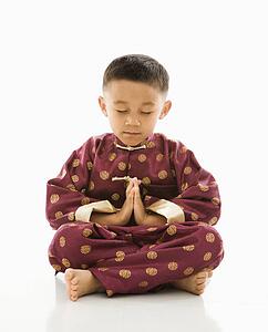 Boy sitting meditating against white background in traditional attire.