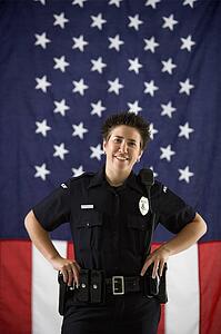 Portrait of mid adult Caucasian policewoman standing with hands on holster and American flag as backdrop smiling at viewer.