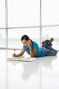 Asian preteen girl lying on floor doing homework while talking on cell phone.