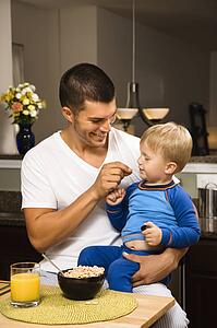 Man feeding toddler son on lap in kitchen 