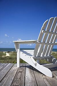 Empty white adirondack chair on wooden deck facing beach.