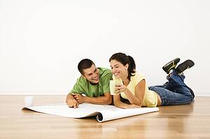 Attractive young adult couple laying on home floor with coffee cups smiling and looking at blueprints