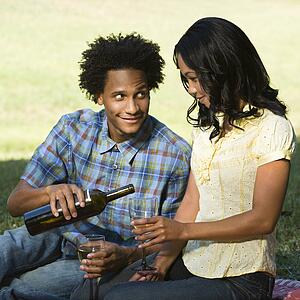 Man pouring woman glass of wine sitting on picnic blanket in park