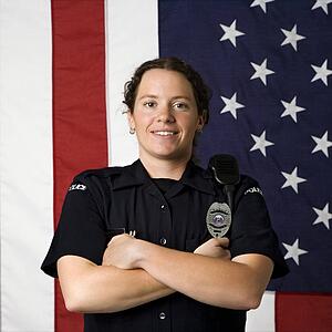 Portrait of mid adult Caucasian policewoman standing with arms crossed with American flag as backdrop smiling at viewer.