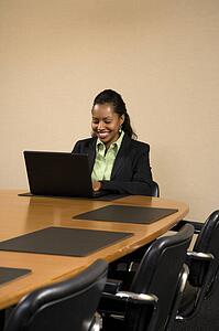 Businesswoman sitting at conference table smiling and typing on laptop computer.