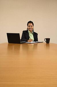 Businesswoman sitting at conference table with laptop computer and coffee cup smiling and writing in book