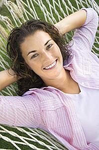 Pretty young adult brunette female laying in hammock with hands behind head smiling at viewer.