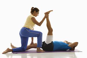 Side view of mid adult woman assisting mid adult man with stretching on exercise mat.