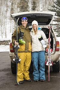 Young couple wearing winter clothes standing by automobile with ski equipment smiling.