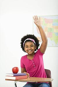 African American girl sitting in school desk raising hand and smiling at viewer.