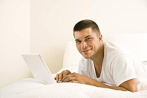 Adult man lying in bed with laptop looking at camera
