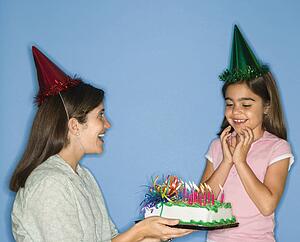 Girl wearing party hat with mother holding birthday cake.