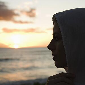 Woman with scarf over head profiled against sunset over ocean