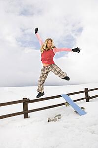 Young woman in winter clothes by fence in snowy field with snowboard jumping into air excitedly