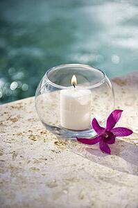 Lit candle in glass bowl with purple orchid next to pool