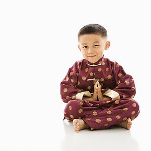 Asian boy sitting meditating against white background in traditional attire.