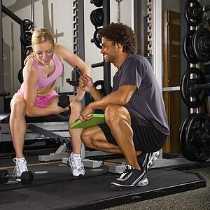 Caucasian mid-adult woman lifting weights while African-American male trainer watches.