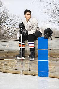 Boy in ice hockey uniform holding hockey stick sitting on sidelines.