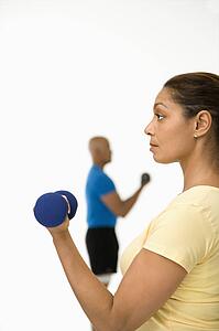 Close up of smiling mid adult woman exercising using dumbbells with mid adult man in background.