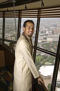 Portrait of young adult male Indian standing near window in tower restaurant smiling at viewer.
