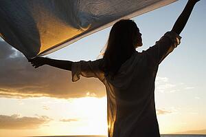 Woman holding up fabric in breeze silhouetted by sunset beside ocean