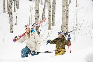Couple walking through snow carrying skis over shoulders smiling