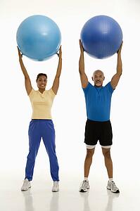 Smiling mid adult man and woman holding blue exercise balls over their heads looking at viewer.