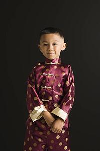 Asian boy in traditional attire standing against black background.