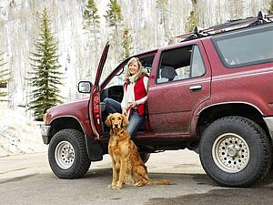 Woman with dog sitting with dirt splattered SUV automobile in snowy rural setting.