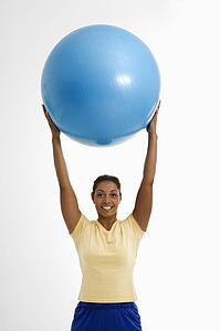 Mid adult woman standing and holding blue exercise ball over her head looking at viewer and smiling.