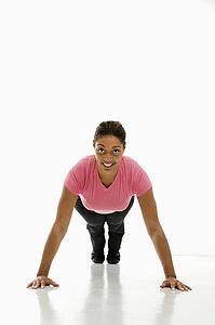 Mid adult woman wearing pink exercise shirt doing pushups while looking at viewer and smiling.