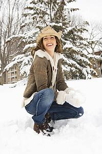 Caucasian young adult female smiling at viewer while kneeling in snow with snowball and wearing straw cowboy hat.