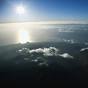 Aerial view of mountainous terrain in Maui, Hawaii with sun shining off the Pacific ocean in background.