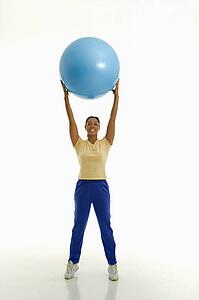 Mid adult woman standing and holding blue exercise ball over her head looking at viewer and smiling.