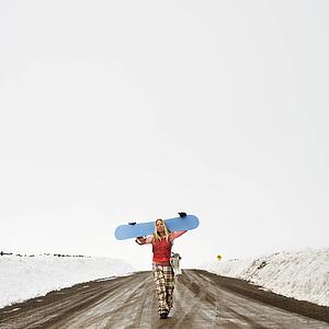 Young woman in winter clothes walking alone down muddy dirt road carrying snowboard and boots
