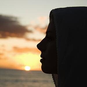 Caucasian mid-adult woman with scarf over head profiled against sunset over ocean.